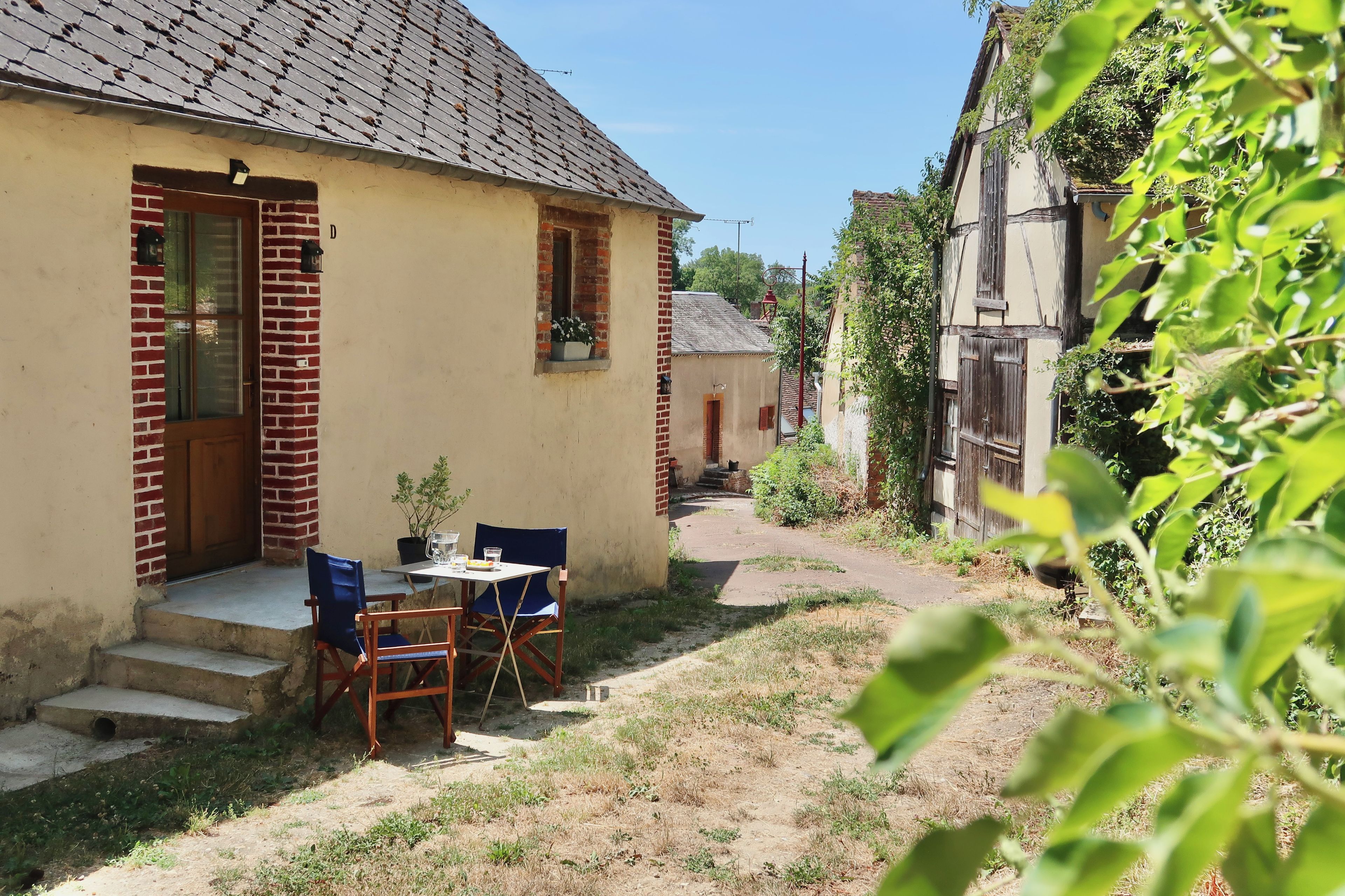 Une petite maison avec une façade en briques, une seule fenêtre ornée d’un pot de fleurs et une table avec deux chaises à l’extérieur.
