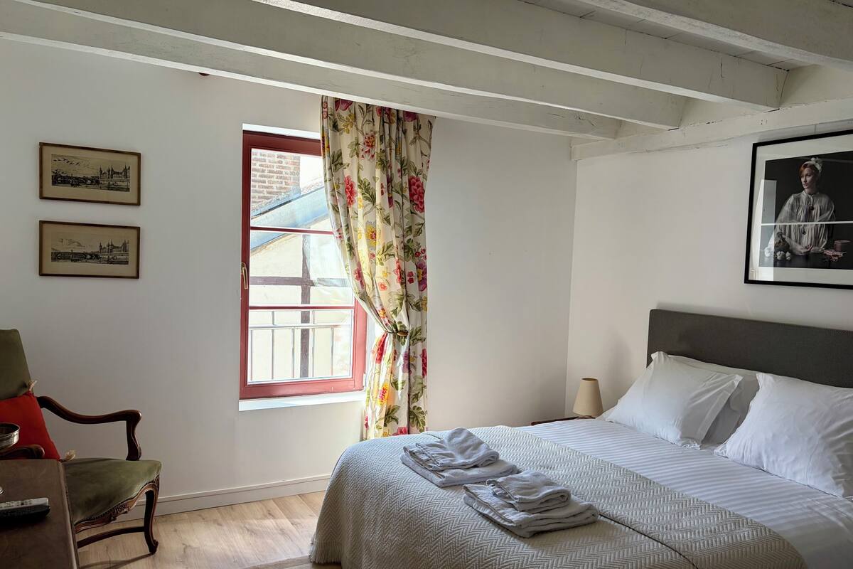 Een slaapkamer met een bed, een stoel, een raam met gordijnen en twee foto’s in lijsten aan de muur.