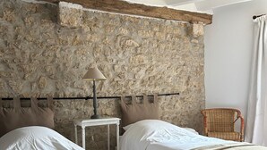 A bedroom with two beds, a stone wall, a small table with a lamp, and a wicker chair. - La grange à grains (Château-Renard)