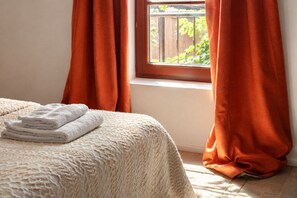 Un lit recouvert d’une courtepointe blanche à motifs, une pile de serviettes blanches, une fenêtre donnant sur un paysage verdoyant et des rideaux orange.