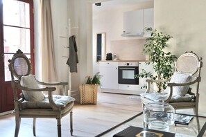 Een kamer met twee antieke stoelen, een glazen bijzettafeltje, een keuken op de achtergrond en een groot raam met gordijnen.