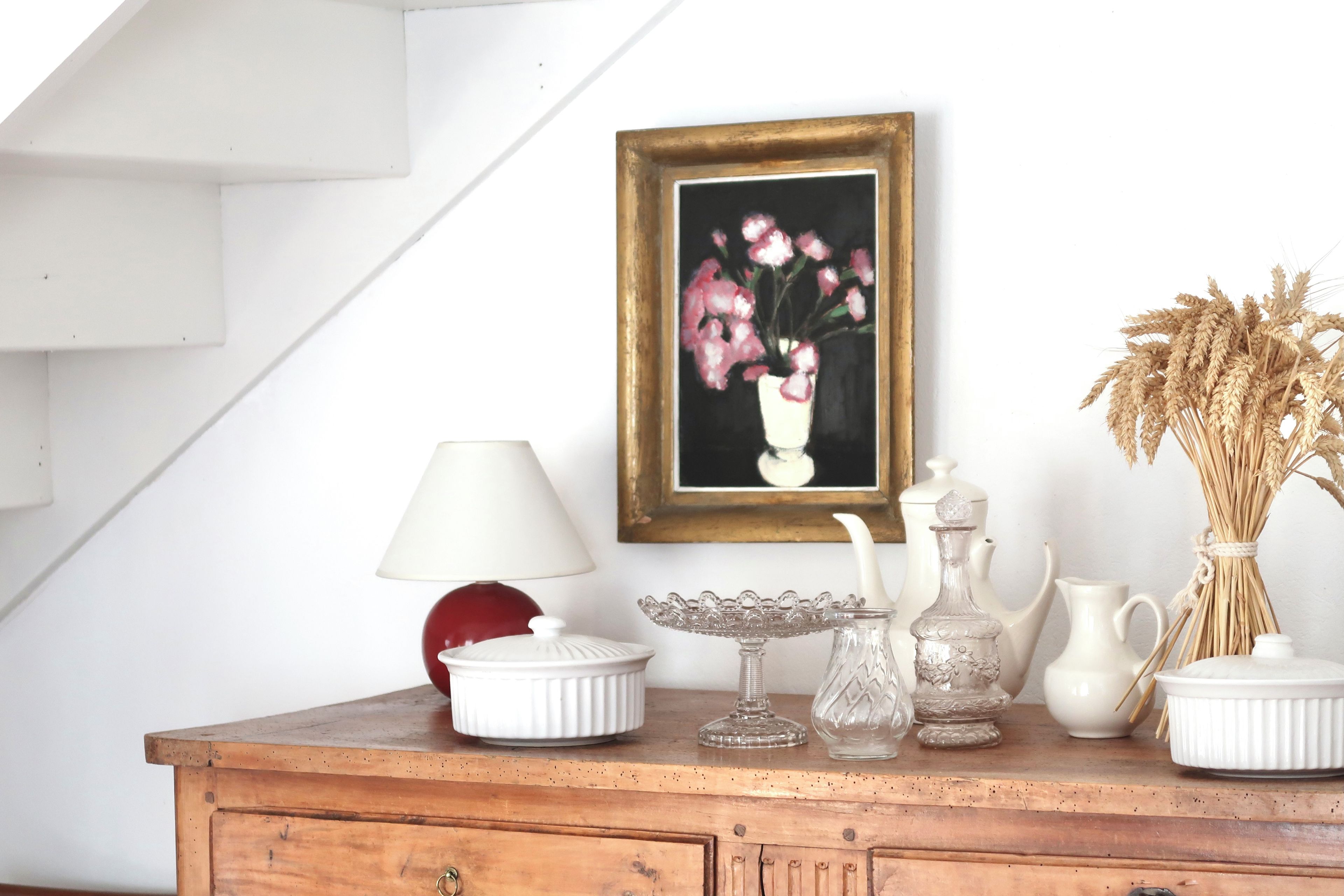Une commode en bois, une lampe blanche, un vase de fleurs et un tableau encadré accroché au mur.
