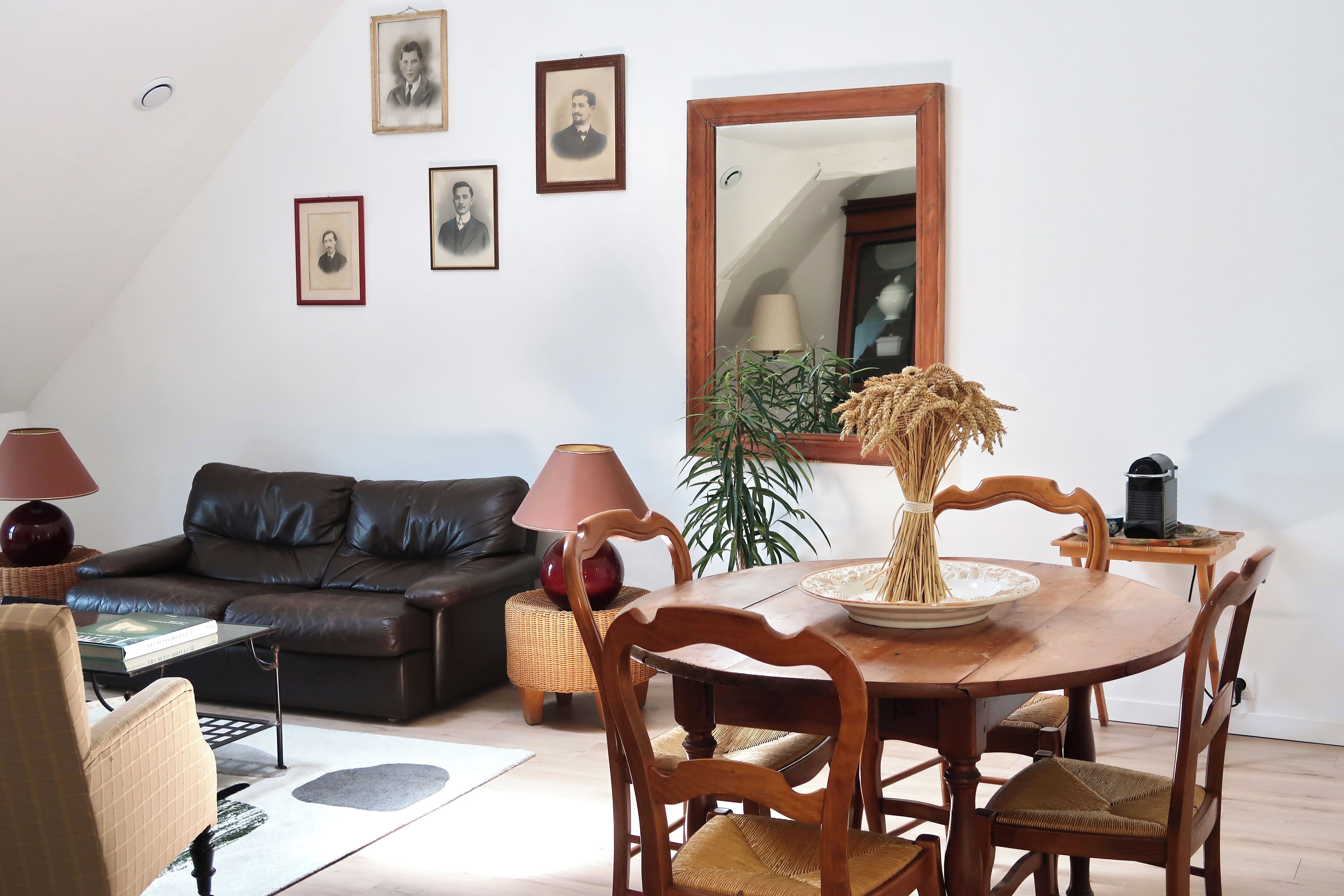 Una sala de estar con un sofá de cuero oscuro, una mesa de comedor de madera con sillas, un espejo grande y cuadros en la pared.