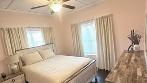 Un dormitorio con cama, mesitas de noche, cómoda, ventilador de techo y ventanas con cortinas.
