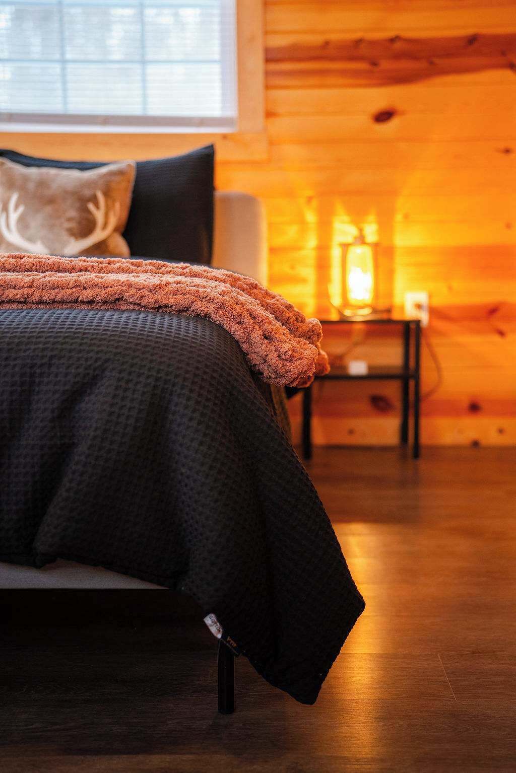 A bed with a dark patterned bedspread and an orange throw blanket, a nightstand with a lamp, and a window with blinds in a wooden room.