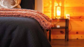 A bed with a dark patterned bedspread and an orange throw blanket, a nightstand with a lamp, and a window with blinds in a wooden room.