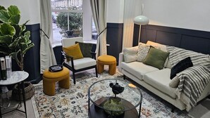 A modern living room with a sofa, armchair, ottoman, and coffee table. There is a large window with curtains, a radiator, and a lamp. - 10% Off Weekly Stay|Chatsworth Retreat|Sleeps 7 (North Yorkshire)
