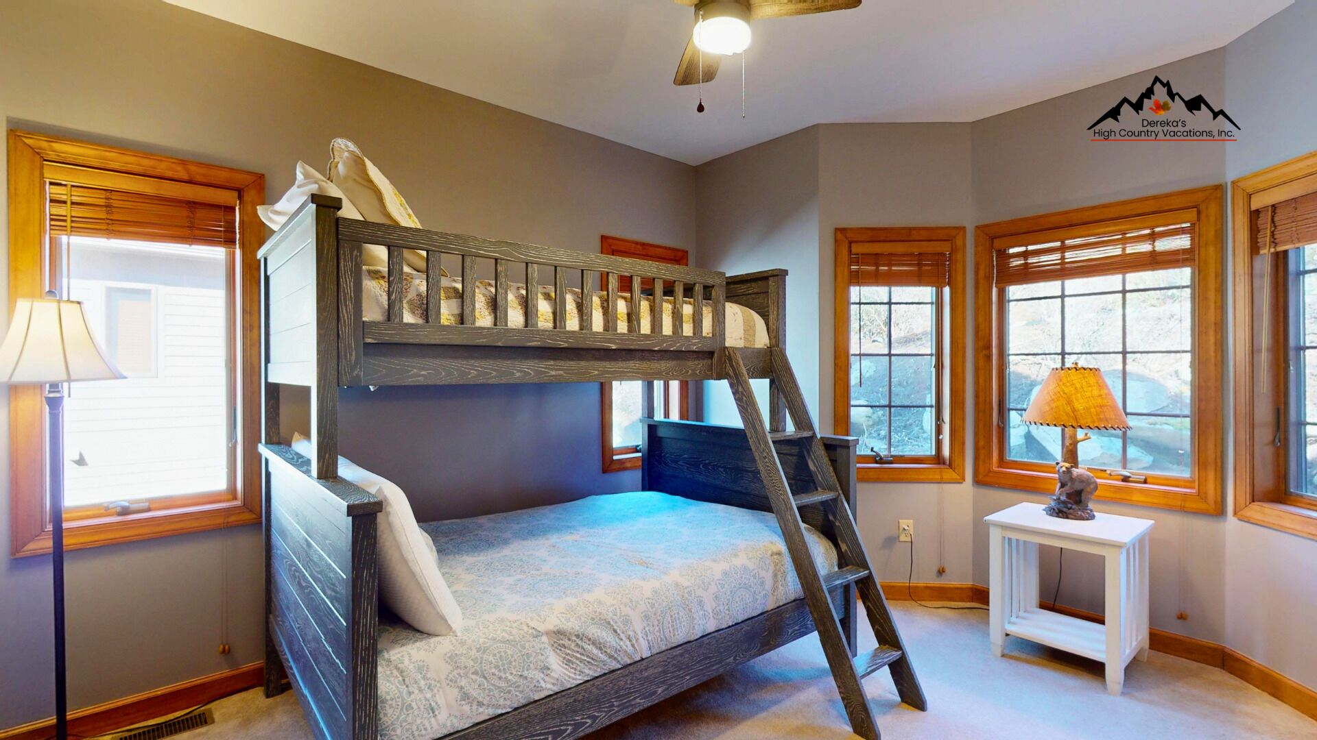 A bunk bed room with a ladder, two bedside lamps, and windows with blinds.