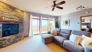 A living room with a stone fireplace, a ceiling fan, and a large window with a view. - Reserve 27D • Big Skyline Views (Sugar Mountain)