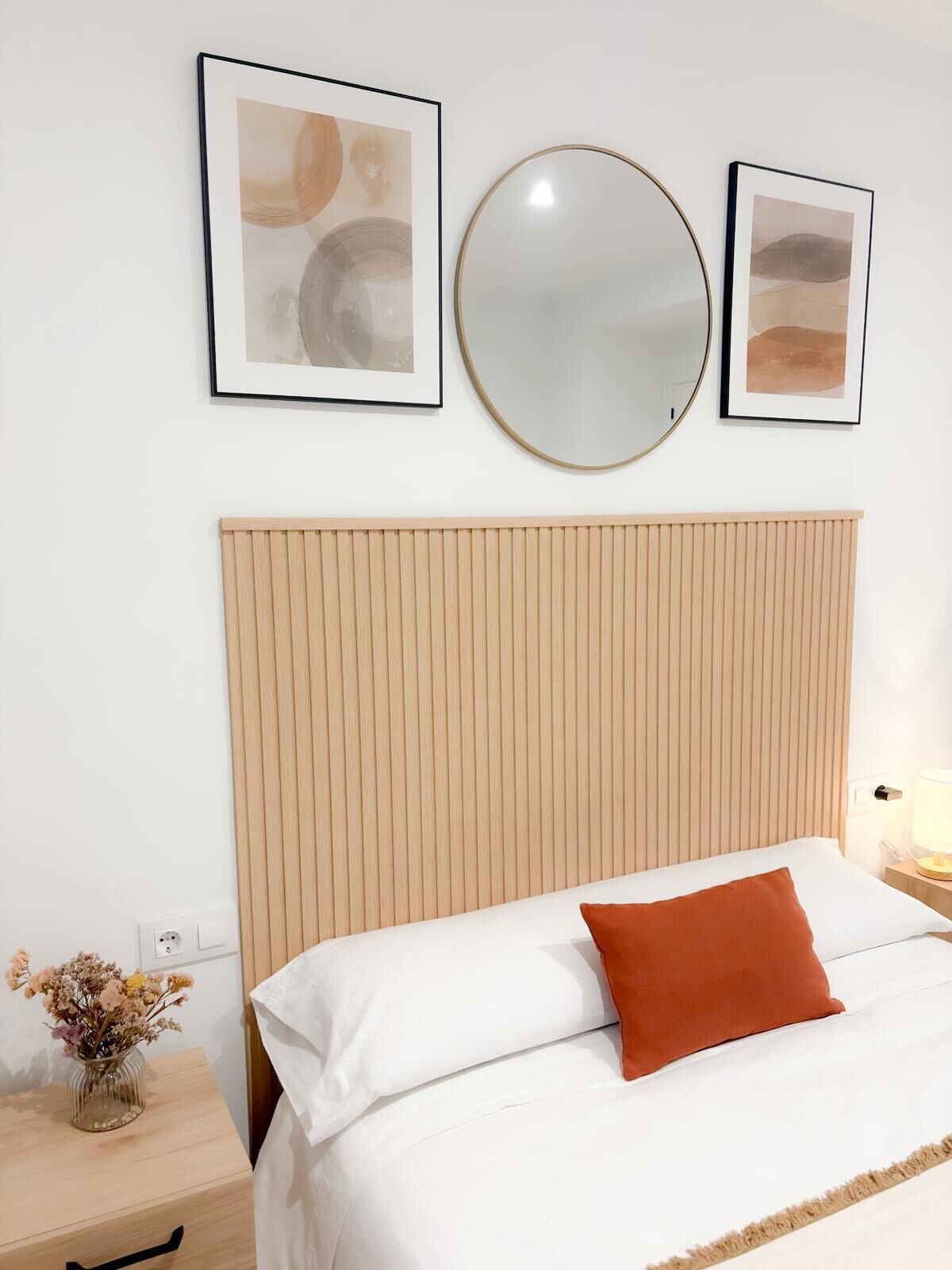 A neatly arranged hotel room with a wooden headboard, a round mirror, and two framed abstract paintings.