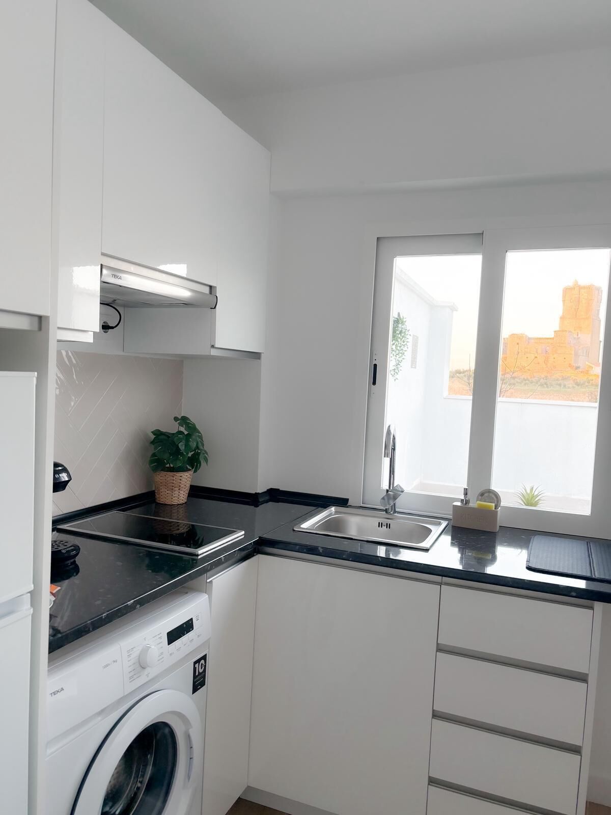 Una cocina moderna con lavadora integrada, un lavamanos y un ventanal con vistas a un edificio lejano.