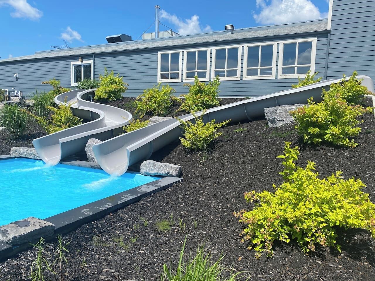 Une piscine dotée de deux toboggans, entourée de parterres aménagés et avec un bâtiment visible au fond.