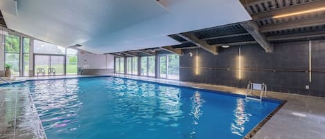 En indendørs swimmingpool med glasvægge, flisegulv og et loft med synlige bjælker.