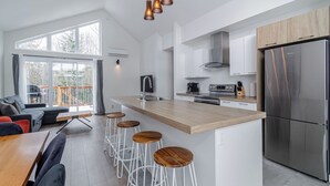 Cozinha moderna com ilha central, banquetas, uma geladeira grande e uma área de jantar com sofá e mesa.