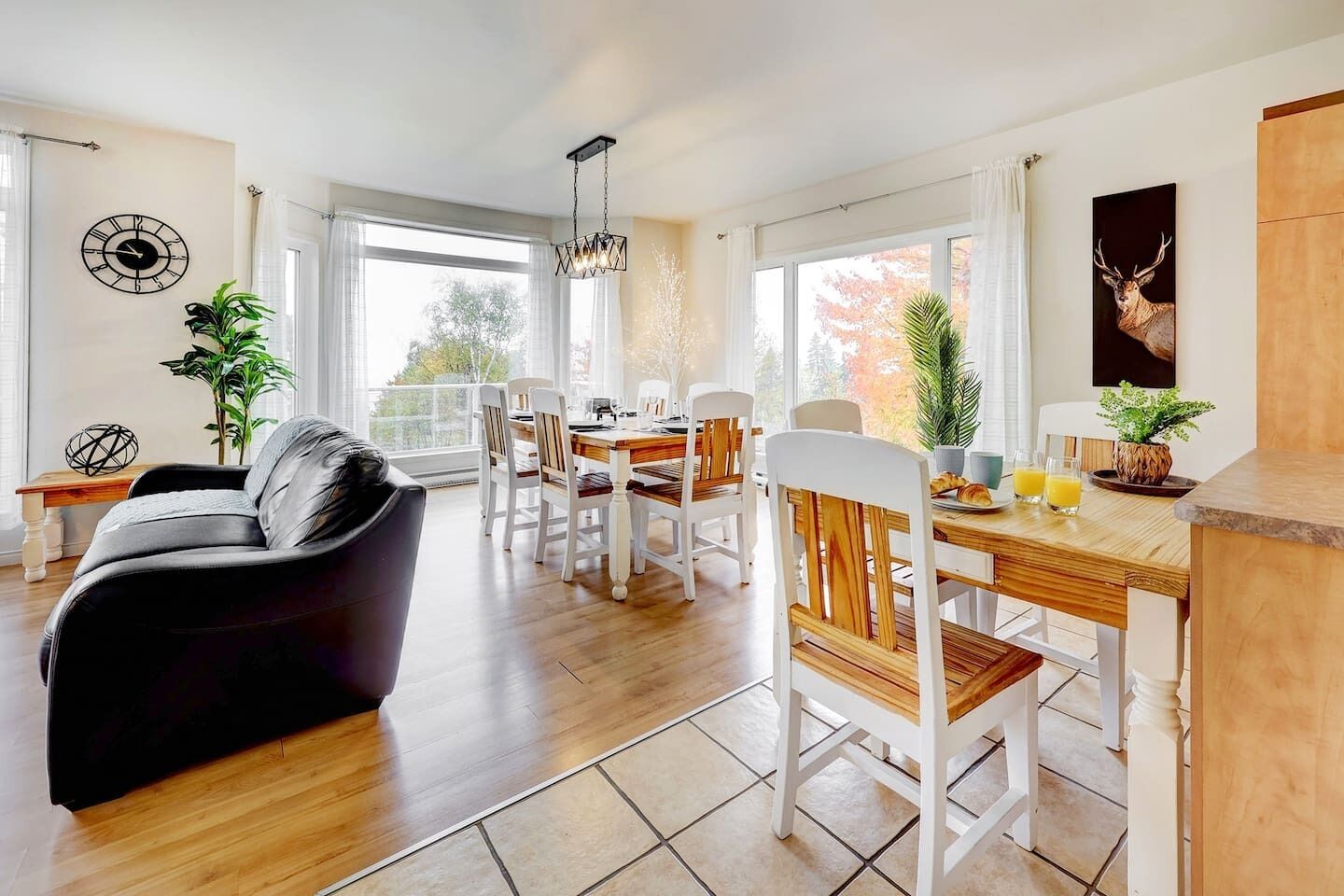 Een moderne woonkamer met een zwarte leren bank, een houten eettafel en witte stoelen. Er is een groot raam met gordijnen, een klok aan de muur en een plant op de tafel.