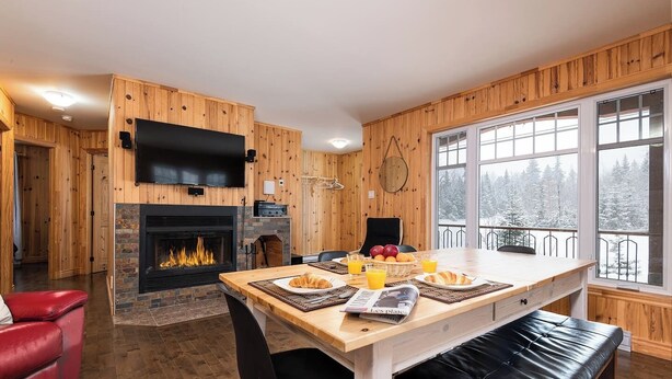 Interior acogedor de una cabaña con chimenea, paredes de madera, zona de comedor con desayuno y un ventanal con vistas a un paisaje nevado.
