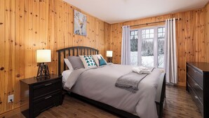 Un dormitorio con paneles de madera, una cama con cabecera, mesitas de noche con lámparas y una ventana con cortinas.