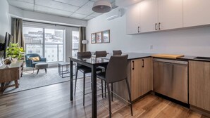 A modern kitchen with wooden cabinets, a dining area with a table and chairs, and a large window offering a city view. - 1CH + Rooftop pool+Cuisine+Smart TV+Gym+750m QC (Québec City)