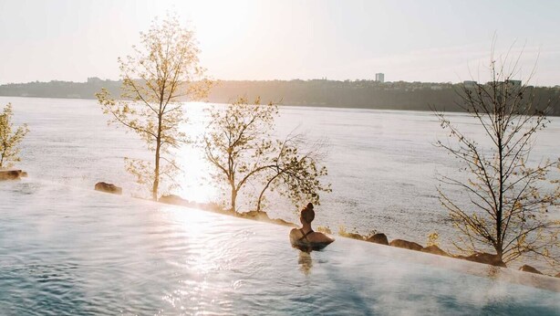 A serene riverside scene with a person sitting on a rock, trees, and a distant city skyline. - 1CH + Rooftop pool+Cuisine+Smart TV+Gym+750m QC (Québec City)