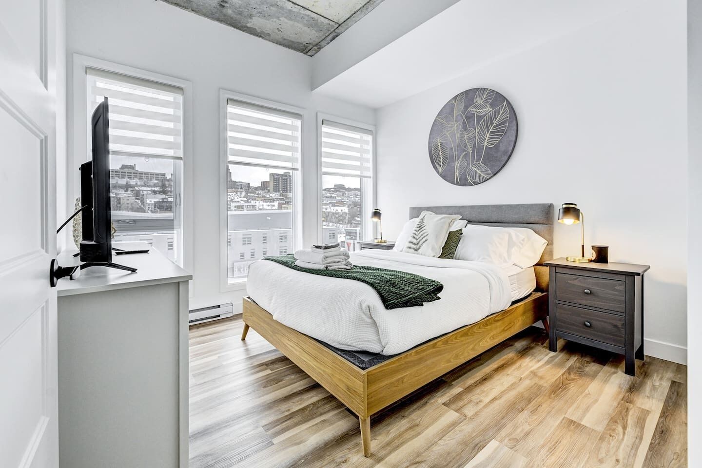 Une chambre moderne avec un lit en bois, un téléviseur posé sur un meuble et une vue sur la ville grâce à de grandes fenêtres.