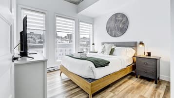 Une chambre moderne avec un lit en bois, un téléviseur posé sur un meuble et une vue sur la ville grâce à de grandes fenêtres.