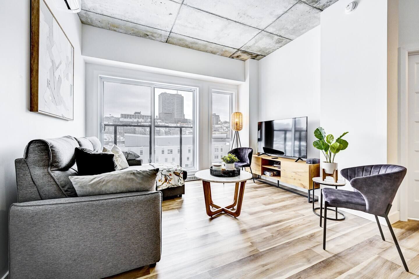 Un salon moderne avec un canapé gris, une table basse, un téléviseur et une grande fenêtre donnant sur la ville.