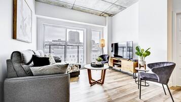 Un salon moderne avec un canapé gris, une table basse, un téléviseur et une grande fenêtre donnant sur la ville.