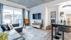 Uma sala de estar moderna com sofá, mesa de centro, mesa de jantar com cadeiras, TV fixada na parede e uma janela grande com cortinas.