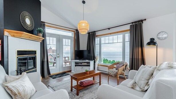 A modern living room with a fireplace, a television, and a large window offering a view of the outdoors. - Panoramic View: 2BR + FP + BBQ + Terrace - Upper (Petite-Rivière-Saint-François)