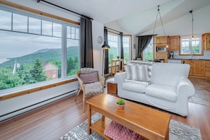 A spacious living room with a large window, a white sofa, a wooden coffee table, and a wicker chair. - Panoramic View: 2BR + FP + BBQ + Terrace - Upper (Petite-Rivière-Saint-François)