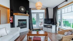 A modern living room with a fireplace, television, and large windows offering a view of the outdoors. - Panoramic View: 2BR + FP + BBQ + Terrace - Upper (Petite-Rivière-Saint-François)