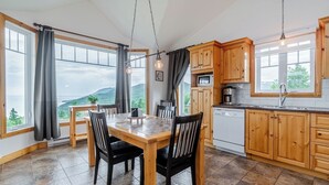 A modern kitchen with wooden cabinets, a dining table with chairs, and a view of the mountains through large windows. - Panoramic View: 2BR + FP + BBQ + Terrace - Upper (Petite-Rivière-Saint-François)