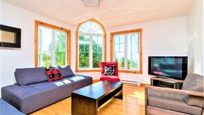 Una sala de estar moderna con un sofá gris, una silla roja, una mesa de centro y un televisor. Amplias ventanas con marcos de madera ofrecen luz natural y vistas al exterior.