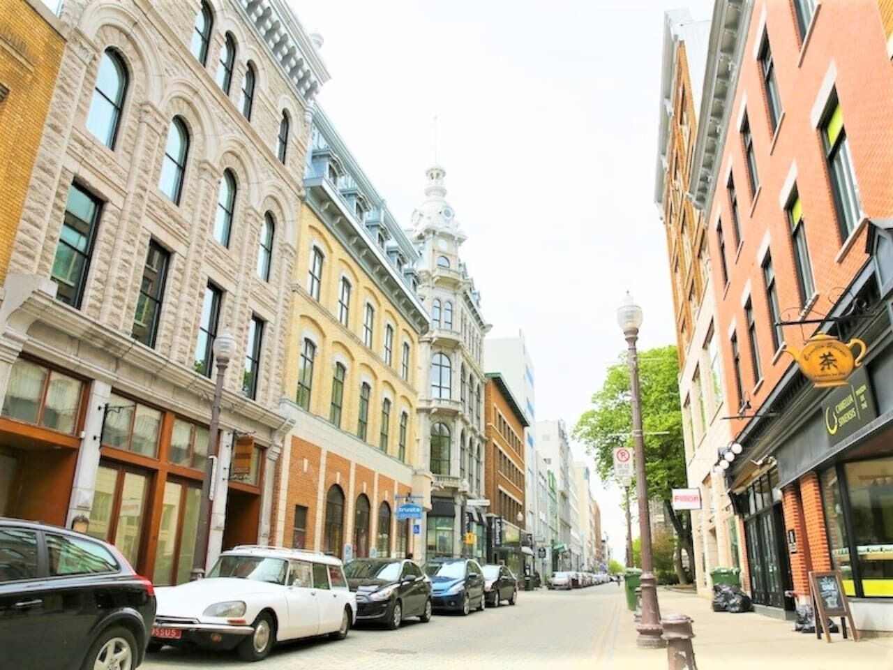 A street view with parked cars, historic buildings, and a restaurant with outdoor seating.