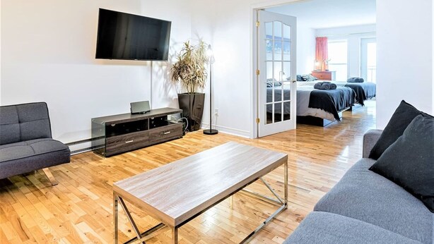 A modern living room with a wooden coffee table, grey sofas, a TV mounted on the wall, and a large potted plant. - Charming Condo - Perfect for Visiting Quebec City (Québec City)