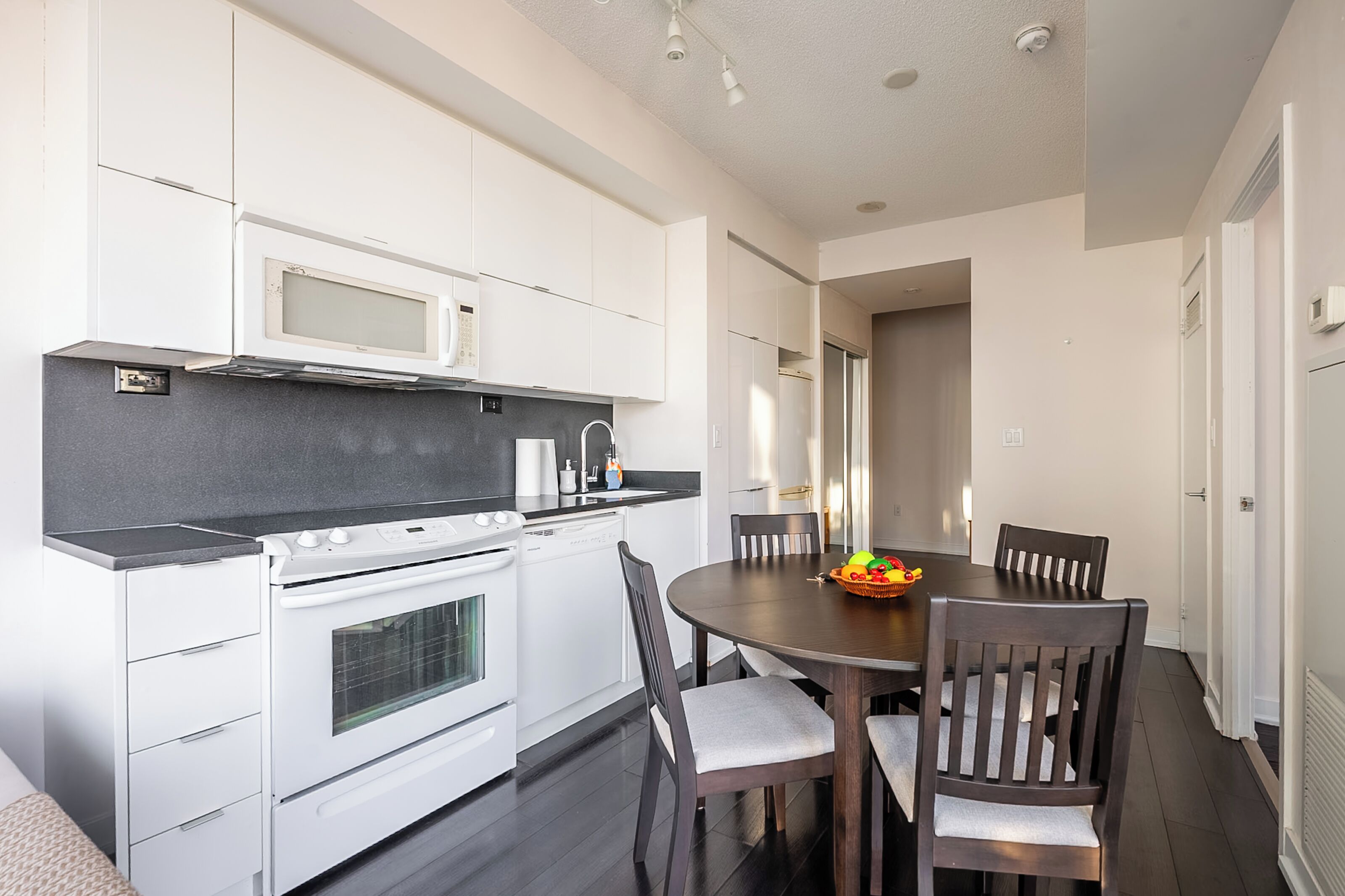 Una cocina moderna con armarios blancos, encimera negra y una zona de comedor con mesa y sillas de madera.