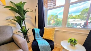 A room with a grey sofa, a black chair with a blue and yellow blanket, a small white table with a green plant, and a large window with a view of buildings and trees.