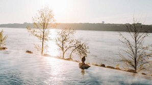 A serene riverside setting with a person sitting on a rock, trees, and a distant city skyline.