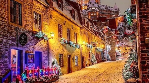 A cobblestone street lined with historic buildings, decorated with festive lights and greenery, leading to a prominent castle-like structure in the background.