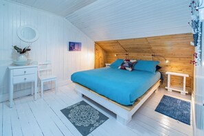 A bedroom with a wooden ceiling, a bed with a blue bedspread, a bedside table, a chair, and a small table with a vase. - Oasis Du Massif - Spa, BBQ, fireplace and nature (Saint-Paul-de-Montminy)
