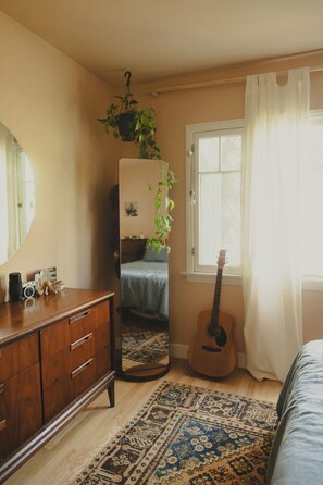Ein Schlafzimmer mit einem Holzkoffer, einem Spiegel, einer Gitarre, einem gemusterten Teppich und einem Fenster mit Vorhängen.