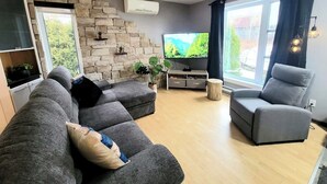 Un salon moderne avec un mur d’accent en pierre, un téléviseur à écran plat, des canapés gris et une grande fenêtre avec des rideaux.