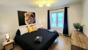 Une chambre à coucher avec un grand lit, une table de chevet, une lampe, une fenêtre avec des rideaux et une plante sur une table en bois.