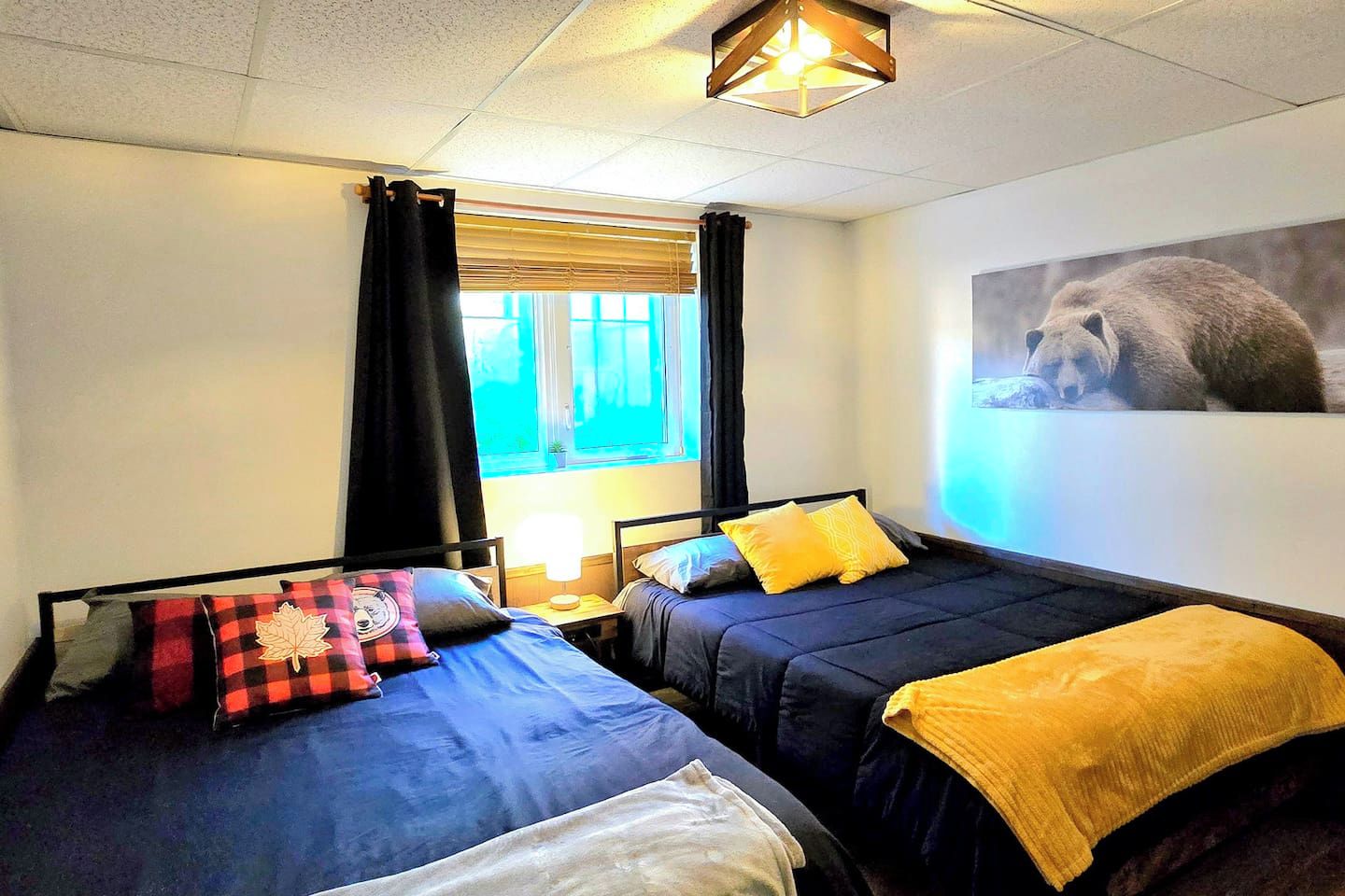 Ein Zimmer mit zwei Betten, einem Vorhangfenster, einer Lampe und einem Bärenbild an der Wand.