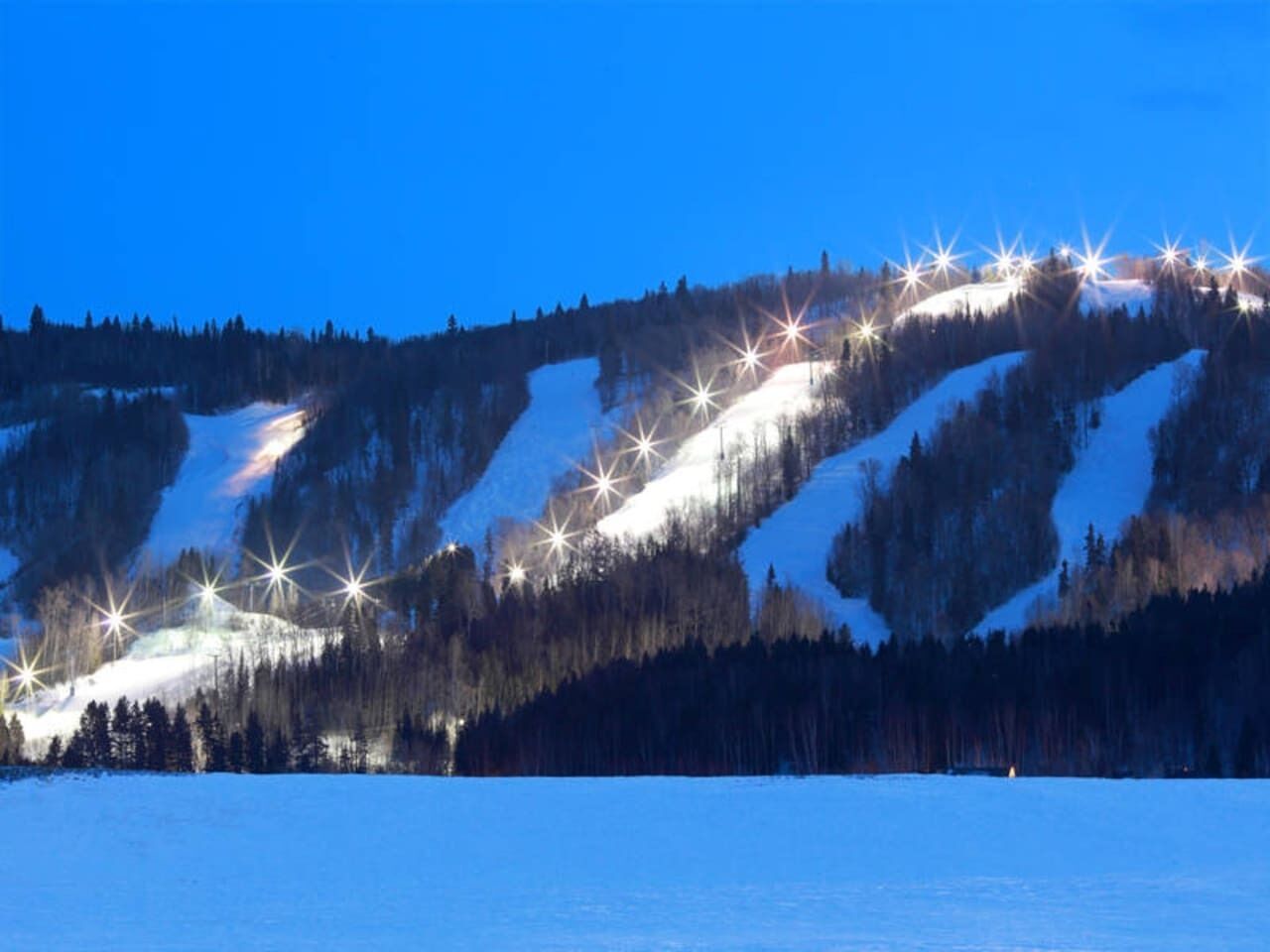 Eine verschneite Berglandschaft im Abenddämmerung mit künstlicher Beleuchtung an den Hängen.
