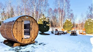 Una estructura de barril de madera con una puerta, rodeada de suelo cubierto de nieve, árboles y asientos exteriores.