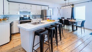 Een moderne keuken met witte kastjes, een centraal kookeiland met krukken en een eethoek met een tafel en stoelen.