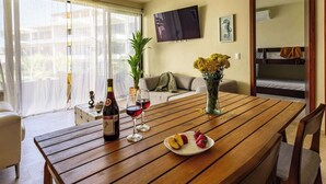 A dining area with a wooden table, two wine glasses, a bottle of wine, a plate with fruit, a vase with flowers, a sofa, a TV, and bunk beds in the background. - Garden-view patio retreat w/ pool & BBQ (Tondoroque)