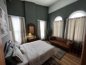 A bedroom with a large bed, a brown sofa, a wooden nightstand, and a mirror.