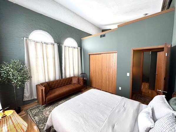 A bedroom with a large bed, a brown sofa, a wooden wardrobe, and a potted plant.